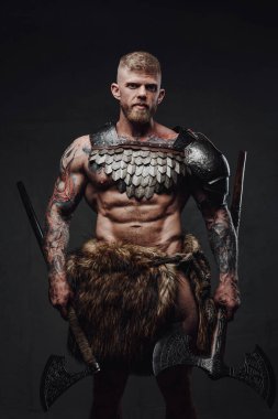 Brutal tattooed warrior wearing light armour and fur holding axes in dark studio clipart