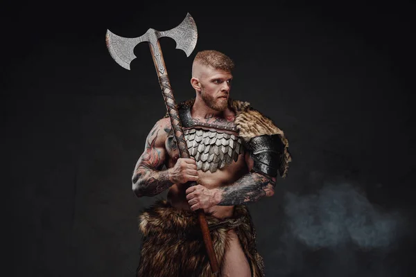 Brutal tattooed warrior wearing light armour and fur holding two-handed axe in dark studio — Stock Photo, Image