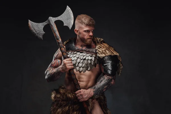 Brutal tattooed warrior wearing light armour and fur holding two-handed axe in dark studio — Stock Photo, Image