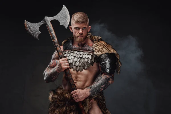 Brutal tattooed warrior wearing light armour and fur holding two-handed axe in dark studio — Stock Photo, Image