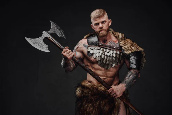 Brutal guerrero tatuado con armadura ligera y piel sosteniendo hacha de dos manos en estudio oscuro — Foto de Stock