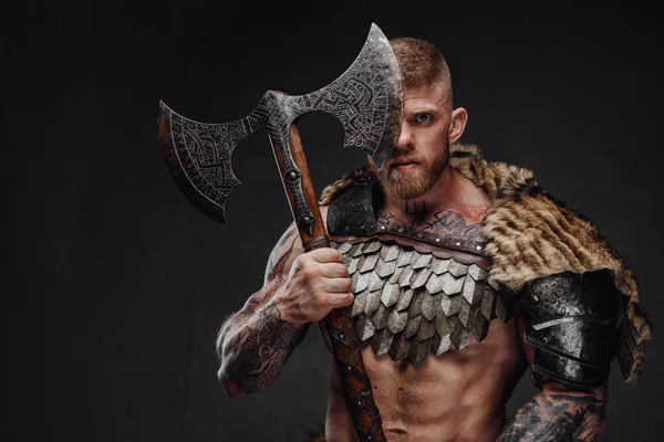 Brutal tattooed warrior wearing light armour and fur holding two-handed axe in dark studio — Stock Photo, Image