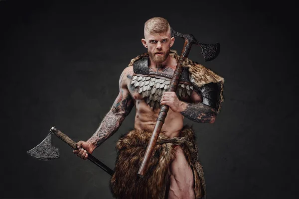 Starker tätowierter Krieger in heller Rüstung und Pelz mit zwei Äxten in Dunkelheit — Stockfoto