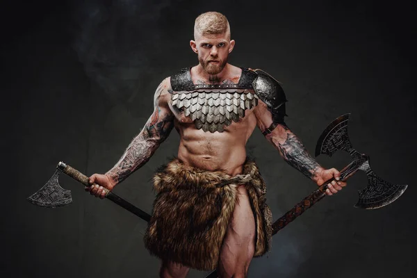 Brutal guerrero tatuado con armadura ligera y hachas de piel en estudio oscuro —  Fotos de Stock