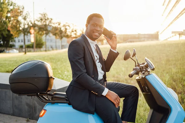 Positivo nero uomo d'affari con scooter utilizzando il suo telefono — Foto Stock
