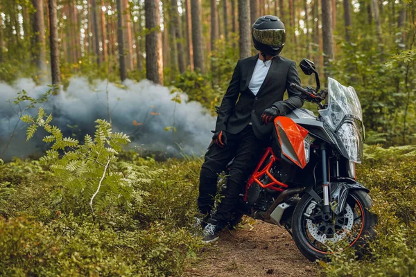 Fresco motociclista in giacca e cravatta in posa con bici contro foresta con fumo — Foto Stock