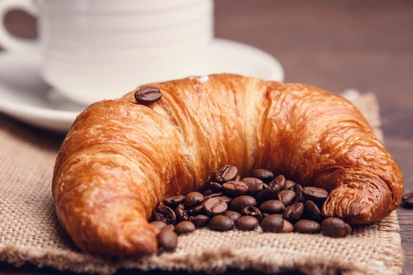 Croissant e grãos de café com uma xícara no fundo — Fotografia de Stock