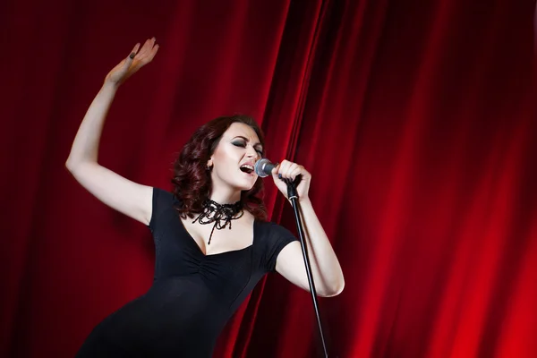 Vackra sjungande flicka på scenen. Skönhet kvinna med mikrofon. Röd gardin bakgrund — Stockfoto