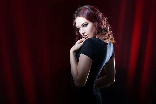 赤いカーテンの背景に黒いドレスの美しいブルネット、魅力的な外観 — ストック写真
