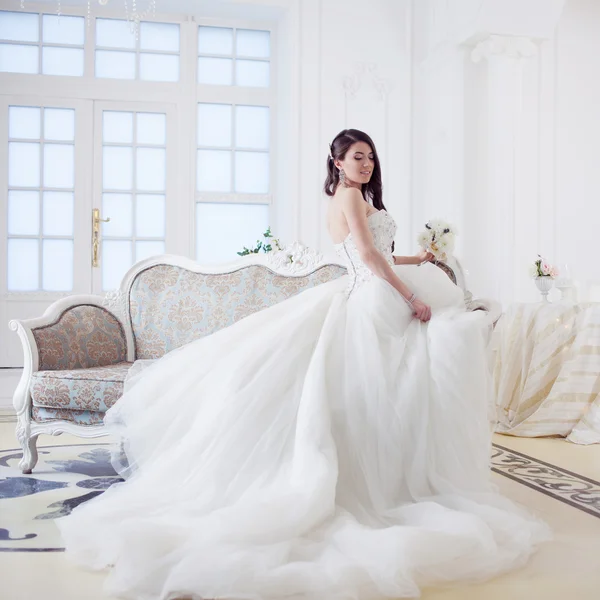 Krásná dívka nevěsta v nádherných šatech sedí na pohovce. Luxusní lehké interiér — Stock fotografie