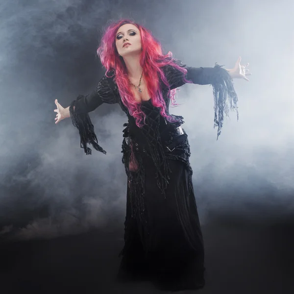 Halloween čarodějnice vytváří kouzlo. Atraktivní žena s červenými vlasy v kostýmu čarodějky stojící nataženou paží, silný vítr — Stock fotografie