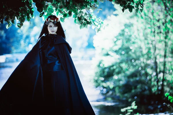 Joven chica de pelo negro en capa larga con una capucha en el bosque oscuro. Efecto de la tonificación — Foto de Stock