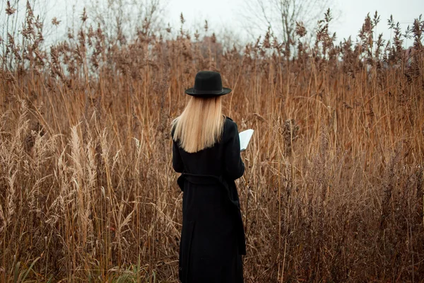 Portré, fiatal, vonzó nő fekete kabátot és kalapot. Shes egy mező olvasó könyv, őszi táj, száraz fű. Nézz vissza — Stock Fotó