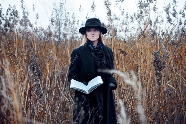 Retrato de mujer joven atractiva en abrigo negro y sombrero. Shes uno en un libro de lectura de campo, paisaje otoñal, hierba seca — Foto de Stock