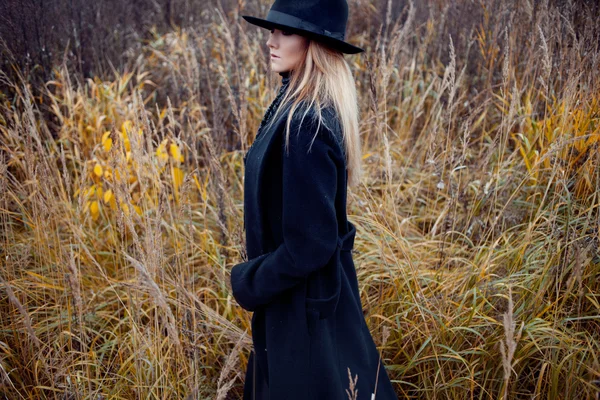 Portret młodej atrakcyjnej kobiety w czarnym płaszczu i kapeluszu. Jesienny krajobraz, sucha trawa — Zdjęcie stockowe