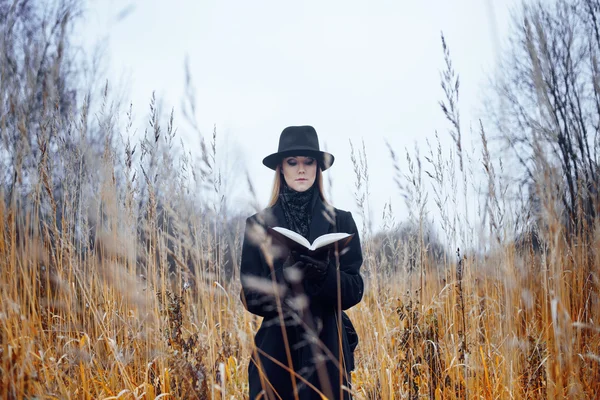 Retrato de mujer joven atractiva en abrigo negro y sombrero. Shes uno en un libro de lectura de campo, paisaje otoñal, hierba seca —  Fotos de Stock