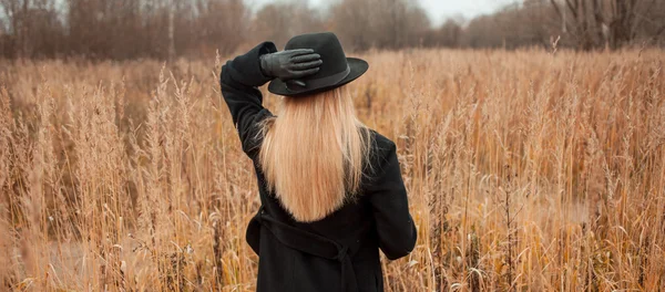 Retrato de mujer joven atractiva en abrigo negro y sombrero. Shes uno en un libro de lectura de campo, paisaje otoñal, hierba seca. Mira atrás. — Foto de Stock