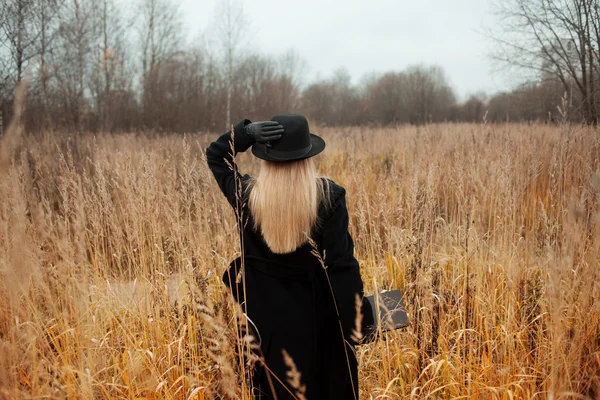 Retrato de mujer joven atractiva en abrigo negro y sombrero. Shes uno en un libro de lectura de campo, paisaje otoñal, hierba seca. Mira atrás. —  Fotos de Stock