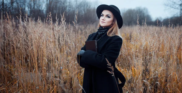 Retrato de mujer joven atractiva en abrigo negro y sombrero. Shes uno en un libro de lectura de campo, paisaje otoñal, hierba seca — Foto de Stock