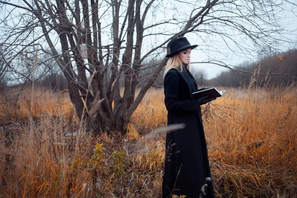 Retrato de mujer joven atractiva en abrigo negro y sombrero. Shes uno en un libro de lectura de campo, paisaje otoñal, hierba seca — Foto de Stock