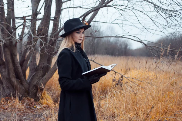 Retrato de mujer joven atractiva en abrigo negro y sombrero. Shes uno en un libro de lectura de campo, paisaje otoñal, hierba seca — Foto de Stock