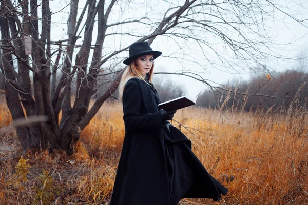 Retrato de mujer joven atractiva en abrigo negro y sombrero. Shes uno en un libro de lectura de campo, paisaje otoñal, hierba seca — Foto de Stock