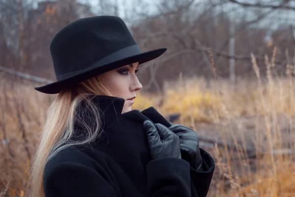 Ritratto di giovane donna attraente in cappotto e cappello nero. Solleva il cappotto collare. Paesaggio autunnale, erba secca — Foto Stock