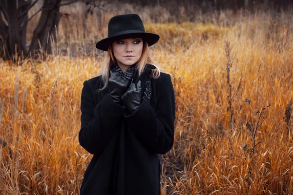 Ritratto di giovane donna attraente in cappotto e cappello nero. Paesaggio autunnale, erba secca — Foto Stock