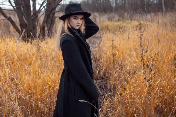 Ritratto di giovane donna attraente in cappotto e cappello nero. Paesaggio autunnale, erba secca — Foto Stock