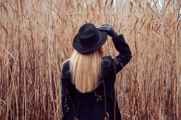 Retrato de mujer joven atractiva en abrigo negro y sombrero. Paisaje otoñal, hierba seca. Mira atrás. Sostener la mano del sombrero —  Fotos de Stock