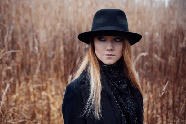 Πορτρέτο της νεαρής ελκυστικής γυναίκας με μαύρο παλτό και καπέλο. Φθινόπωρο τοπίο, ξηρό γρασίδι — Φωτογραφία Αρχείου
