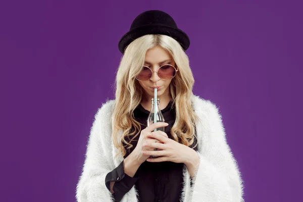 Young beautiful blonde woman in a fashionable hat. Drinking soda from glass bottles. Purple background — Stock fotografie