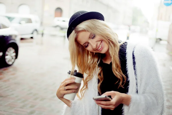 Vrolijke vrouw in de straat drinken koffie in de ochtend en haar smartphone gebruiken. — Stockfoto