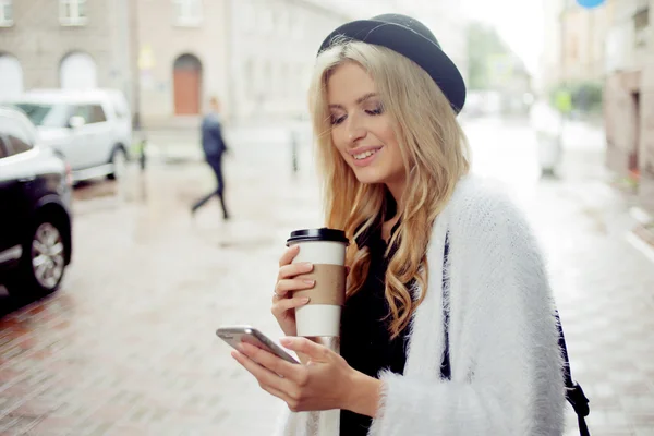 Vrolijke vrouw in de straat drinken koffie in de ochtend en haar smartphone gebruiken. — Stockfoto