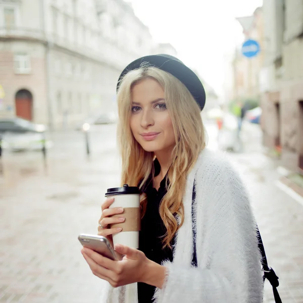 Fröhliche Frau auf der Straße trinkt Morgenkaffee und benutzt ihr Smartphone. — Stockfoto