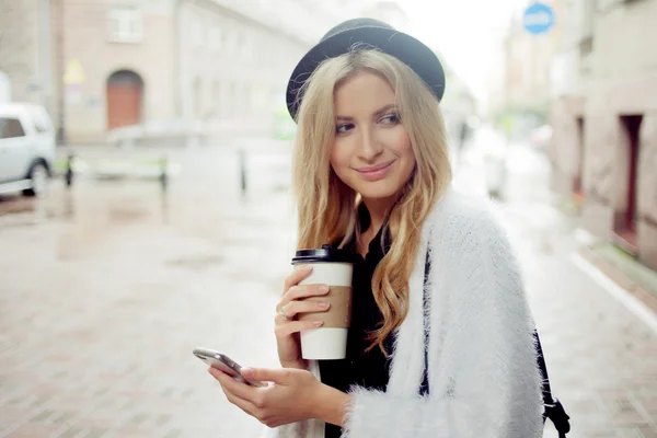 Fröhliche Frau auf der Straße trinkt Morgenkaffee und benutzt ihr Smartphone. — Stockfoto