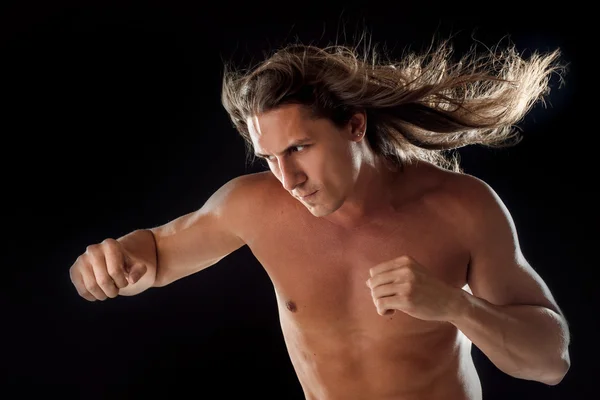 Muscular man with long hair, clasps hands in fist, black background — Stock Photo, Image