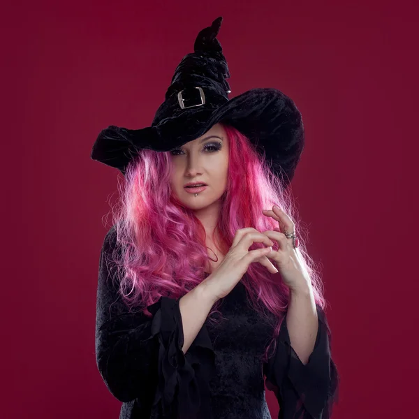 Attraktive Frau mit Hexenhut und Kostüm mit roten Haaren zaubert auf rosa Hintergrund. Halloween, Horror-Thema. — Stockfoto