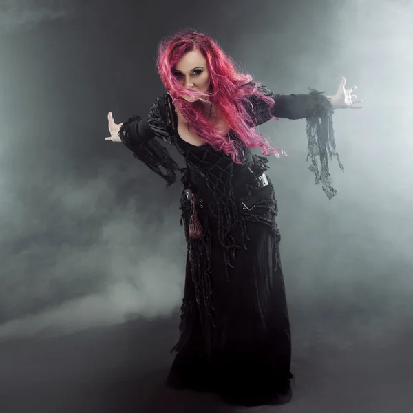 Halloween čarodějnice vytváří kouzlo. Atraktivní žena s červenými vlasy v kostýmu čarodějky stojící nataženou paží, silný vítr — Stock fotografie