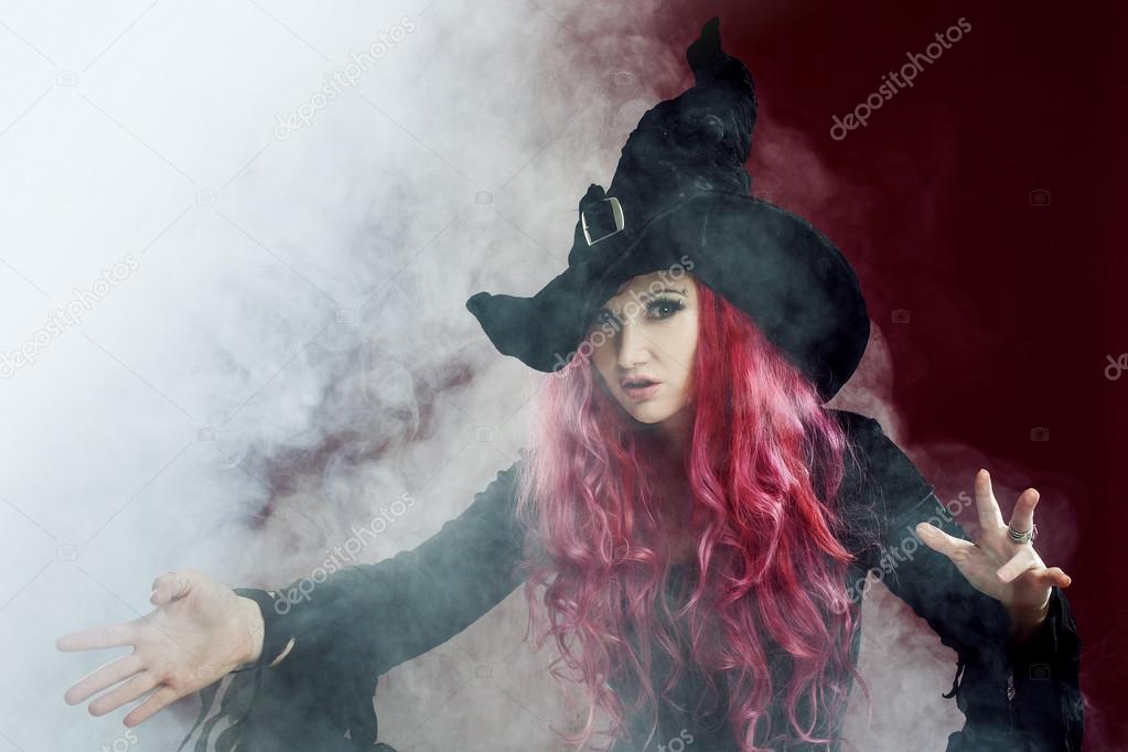Attractive woman in witches hat with red hair performs magic. Smoke and witchcraft