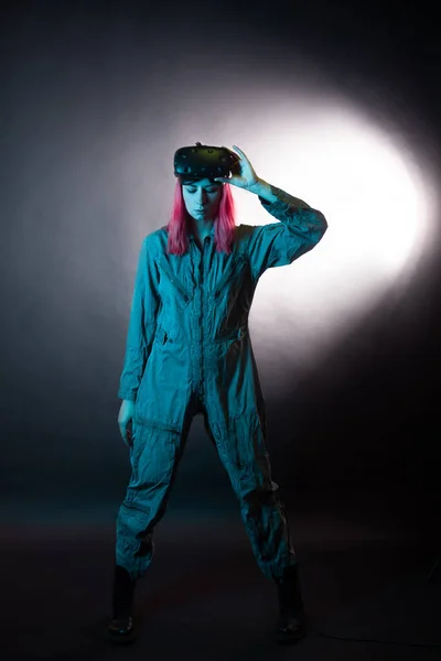 Mujer joven con el pelo rosa y en un mono utiliza gafas de realidad virtual, está en el juego, —  Fotos de Stock