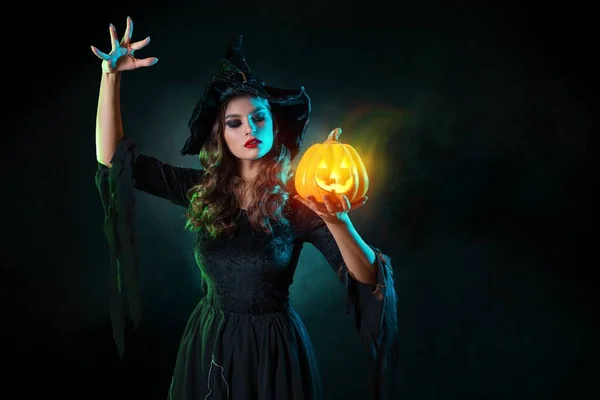 beautiful witch with a hat conjures over jack-o-lantern pumpkin.