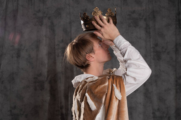 A young king in a fur robe crowns himself, a man holds a crown over his head.