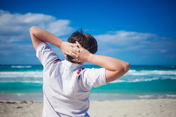 Guapo joven contra el fondo de la playa, relaja sus manos detrás de la cabeza —  Fotos de Stock