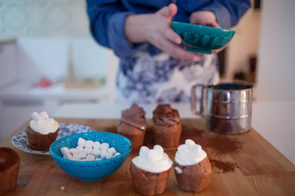 Девушка украшает кексы, держа тарелку, кексы и тарелку ингредиентов для украшения на столе — стоковое фото