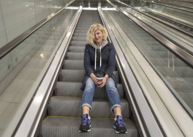 Sarışın kız yürüyen merdivenlerde oturuyor.