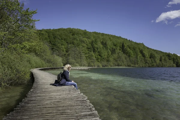 Blondýnka je sezení na mostě poblíž jezera s ohromující modrá voda — Stock fotografie