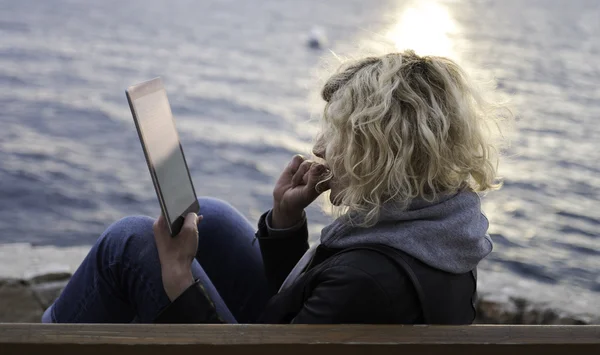 Menina loira com cabelo encaracolado está sentado no banco e absorbedly assistindo algo em tablet — Fotografia de Stock