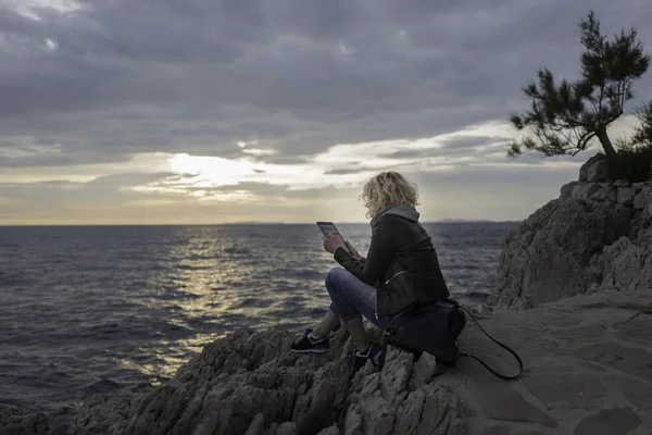 Hermosa chica rubia está sentada en las piedras cerca del mar y mirando la tableta — Foto de Stock