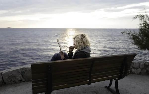 Blonde fille avec les cheveux bouclés est assis sur le banc et regarder absorbant quelque chose dans la tablette — Photo